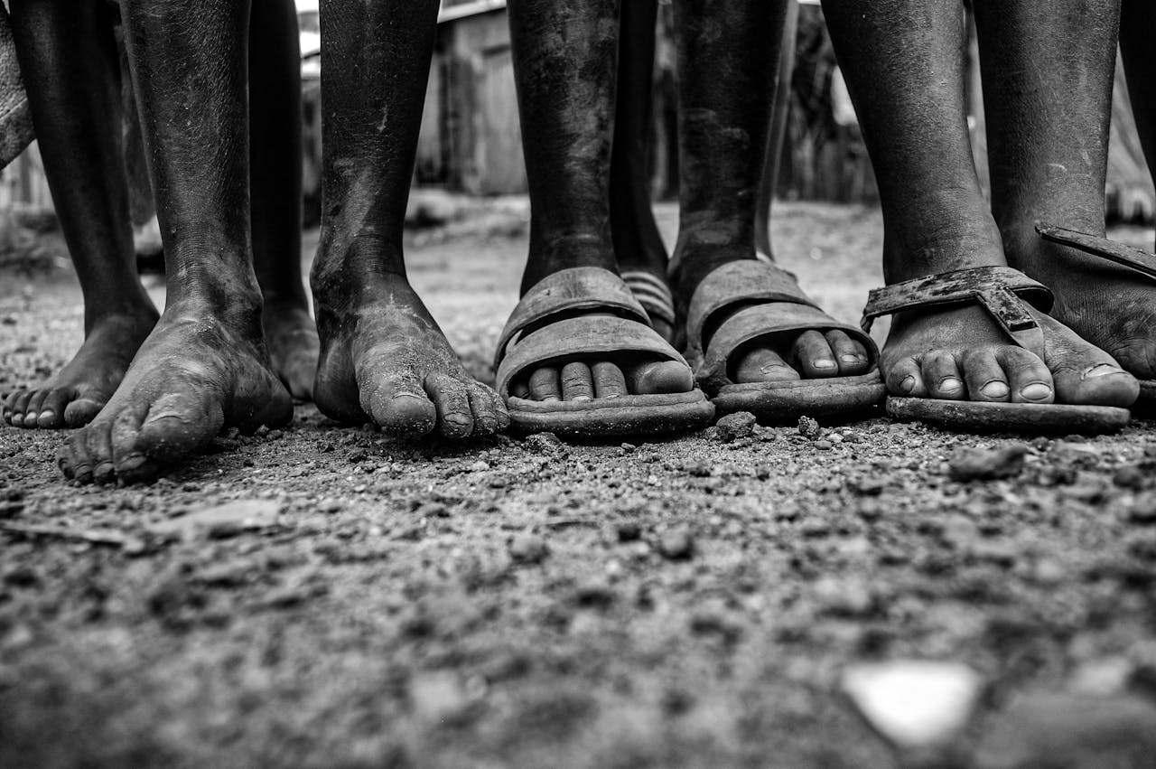Close-up of children's feet in sandals and barefoot, standing on dusty ground.