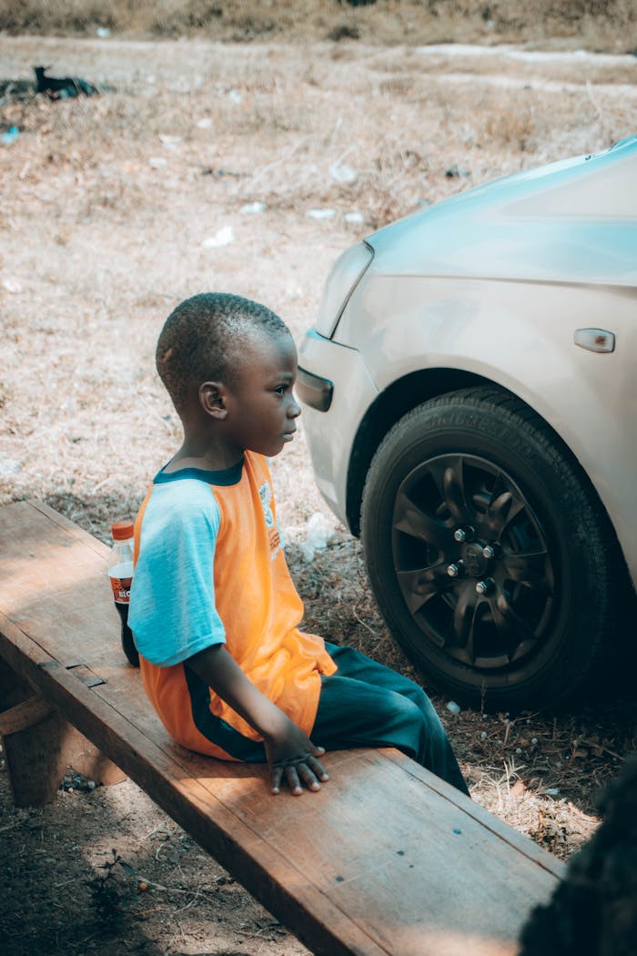 about-07 A young boy sits thoughtfully on a bench beside a car in a sunny outdoor setting.