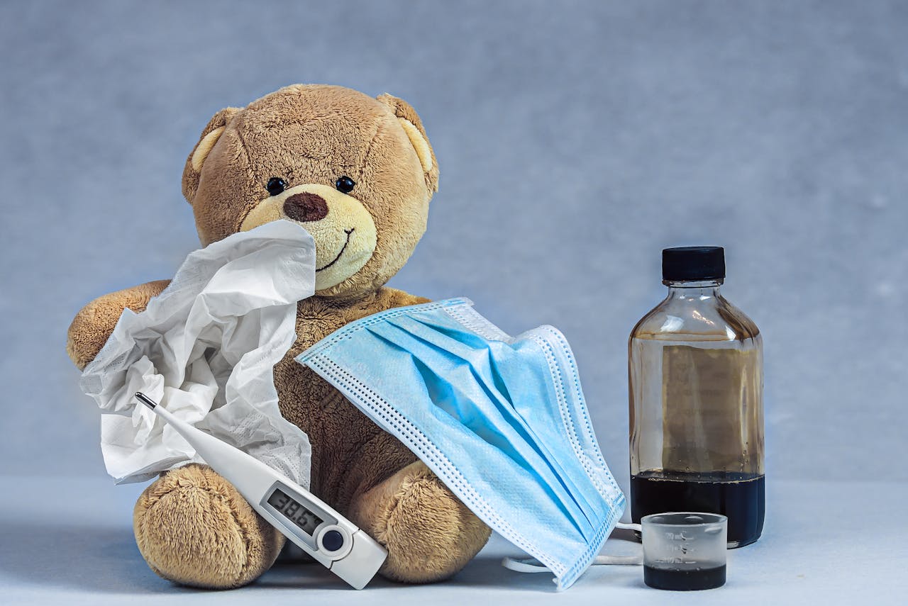 A teddy bear with cough syrup, a mask, thermometer, and tissue highlights an illness concept.
