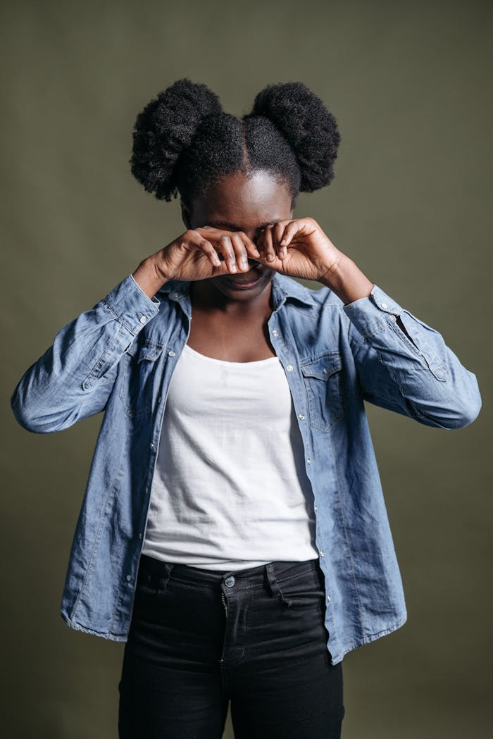 A woman expressing sadness, wiping tears, wearing casual attire against a neutral background.